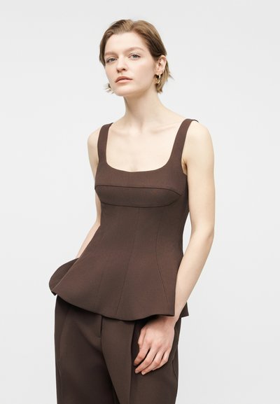 Νέα γυναίκα με κοντά μαλλιά φοράει καφέ αμάνικη μπλούζα με πεπλουμ και ταιριαστά παντελόνια, ποζάροντας με το ένα χέρι στην τσέπη μπροστά σε λευκό φόντο.