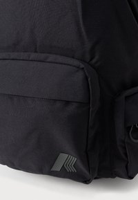 Mochila negra hecha de un tejido duradero, con un bolsillo frontal con solapa y un logo en material de goma gris. Superficie texturizada.