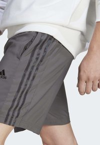 Grijze sportshorts gemaakt van lichte stof met een ademende textuur. Bevat drie zwarte strepen en een adidas-logo aan de zijkant.
