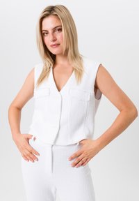 Witte mouwloze vest met een V-hals, textuurstof, twee voorzakken en knoopsluiting, gecombineerd met bijpassende witte broek.