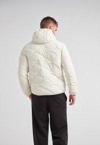 Vadderad vit jacka med huva, med en quiltad design med vågmönster. Bärs med svarta byxor, visad bakifrån.