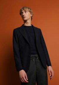 Blazer azul marino ajustado con dos bolsillos frontales, debajo un cuello de tortuga negro, combinado con pantalones grises con cordón sobre un fondo naranja.