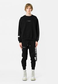 Sudadera negra con cuello redondo y etiqueta frontal, combinada con pantalones jogger negros que presentan cremalleras y detalles desgastados. Botas blancas.