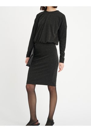 Nainen pukeutuneena mustaan, vartalonmyötäiseen, kimaltavaan mekkoon, jossa on pitkät hihat, läpikuultaviin mustiin sukkahousuihin, mustiin slingback-korkoihin ja mustaa clutch-laukkua kädessään.