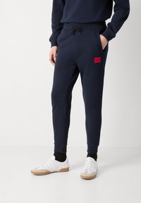 Marineblauwe joggingbroek met elastische tailleband en trekkoord, voorzien van een rood logo-patch, gecombineerd met witte sneakers en zwarte sokken.