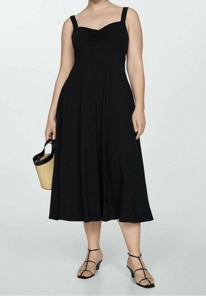 Woman wearing a sleeveless black midi dress with gathered bust, holding straw handbag, and black strappy low-heel sandals.