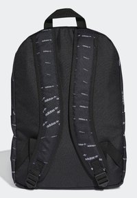 Svart Adidas ryggsäck med justerbara vadderade axelremmar som har upprepade vita Adidas-logotyper och ett handtag på toppen.