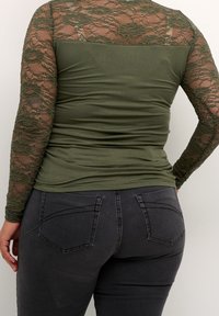 Oliivinvihreä pusero, jossa pitkät pitsihihat ja kukkakuvioitu tekstuuri; vartalonmyötäinen malli, yhdistetty tummanharmaiden farkkujen kanssa.