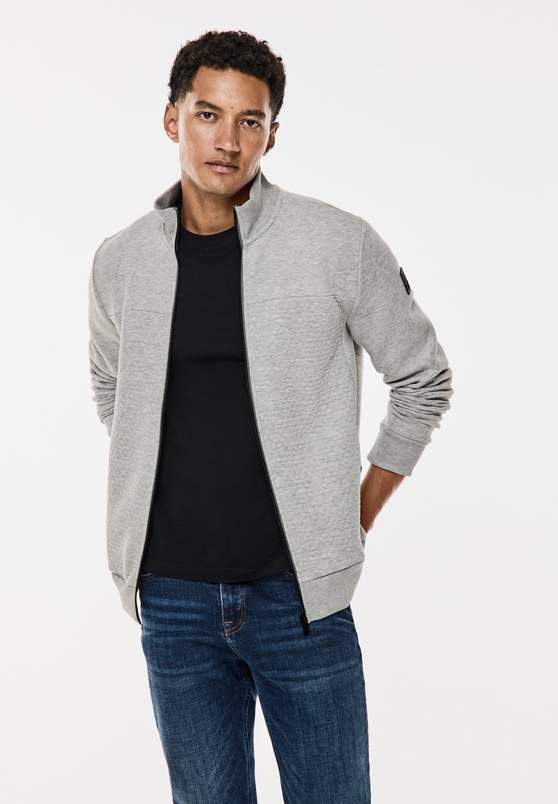 Gray zip-up jacket with a textured pattern, standing collar, two front pockets, worn over a black t-shirt and blue jeans.