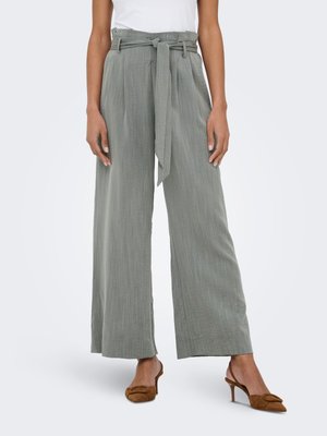 Vrouw draagt grijze, hoog taille, wijde broek met een stoffen riem gestrikt in de taille, gecombineerd met bruine slingback schoenen met hak.