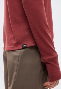 Punane pika varrukaga särk sõrmusega avaga ja väikese musta logomärgiga, kantud pruuni püksiga, näidates käsivart ja puusa külgvaates.