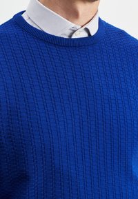 Neulottu sininen villapaita, jossa on teksturoitu, resorikuosinen pinta. Pyöreä kaula-aukko, jossa näkyy harmaa kauluspaita alla.