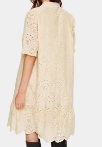 Vaaleanbeige puuvillamekko, jossa on brodeerattuja leikattuja yksityiskohtia, lyhyet hihat röyhelöpuvuilla ja kerroksellinen helmala.