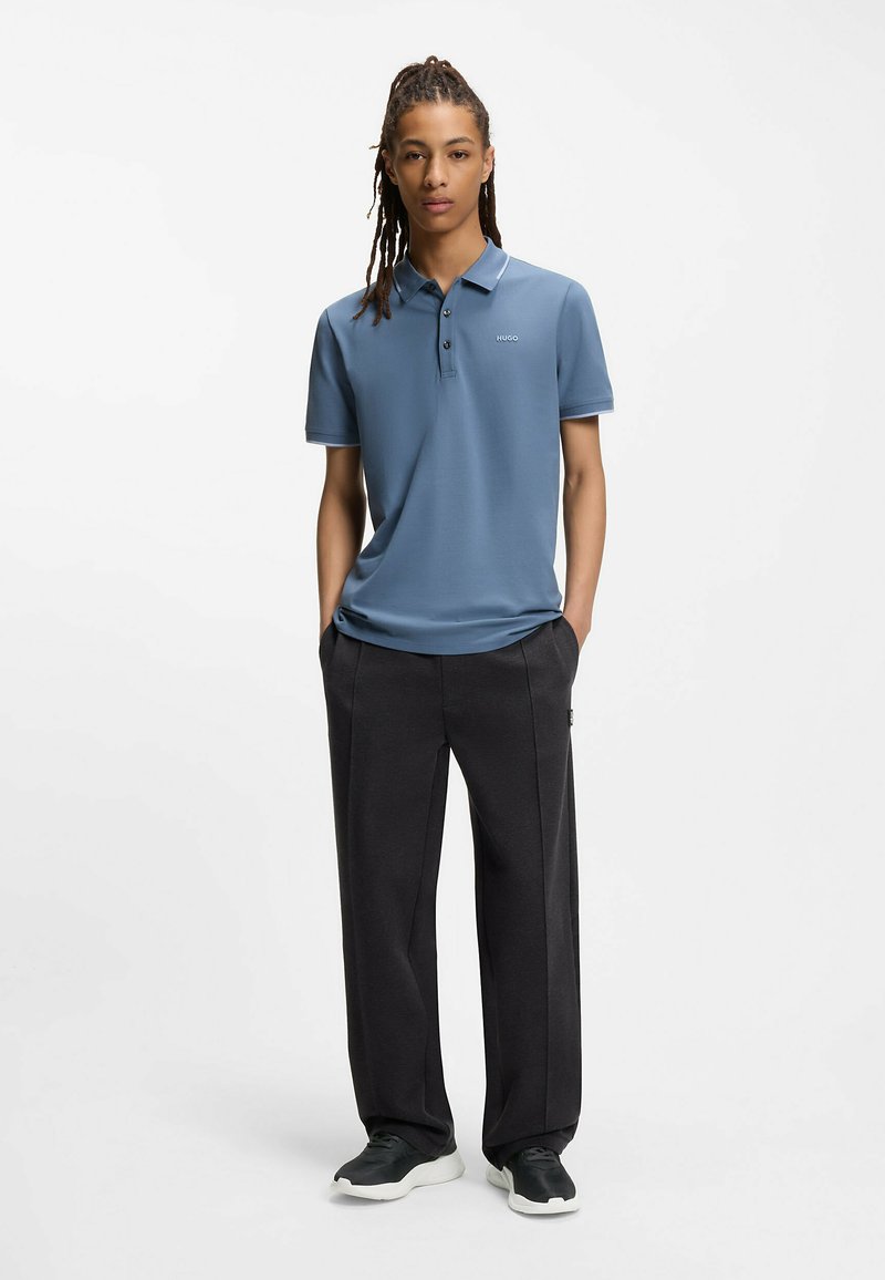 Hellblaues Poloshirt mit einer Knopfleiste mit drei Knöpfen, versehen mit einem kleinen Logo. Kombiniert mit dunkelgrauen, weit geschnittenen Hosen und schwarzen Sneakern.