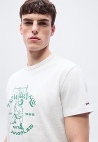 Νεαρός άνδρας με κοντά καστανά μαλλιά, που φοράει λευκό μπλουζάκι με πράσινο γράφημα "Tommy's Gym Los Angeles 1985" σε απλό φωτεινό φόντο.