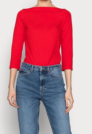 Maglietta rossa a maniche lunghe con scollatura quadrata, abbinata a jeans blu di denim a vita alta. Il tessuto appare morbido e con una texture liscia.