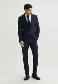 Granatowy, dopasowany garnitur z jednorzędową marynarką, klapami w kształcie litery notch oraz dopasowanymi spodniami. Biała koszula i ciemny krawat dopełniają całość. Czarne buty.