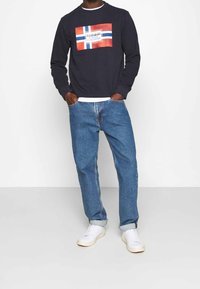 Granatowy sweter z grafiką przedstawiającą czerwono-niebieski wzór norweskiej flagi, zestawiony z jasnoniebieskimi dżinsami i białymi trampkami.