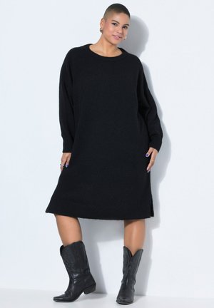 Person med kort hår iført en sort langærmet sweaterkjole og sorte læderstøvler til midt på læggen, der står mod en hvid baggrund.