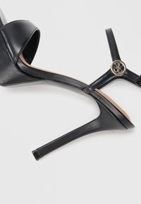 Sandali neri con tacco alto, punta chiusa e cinturino alla caviglia. Materiale in pelle liscia, tacco stiletto sottile e dettaglio decorativo con logo argentato.