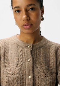Gebreide cardigan in lichtbruin met kabelpatronen, grote parelknopen en een ronde halslijn, met een gestructureerde, cozy uitstraling.
