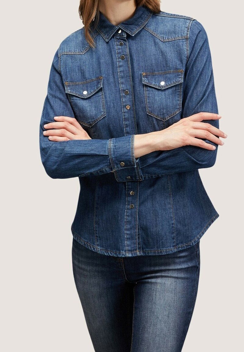 Chemise en denim à manches longues, de couleur bleu clair, avec fermeture boutonnée sur le devant, deux poches poitrine et un empiècement de style western. Texture lisse.