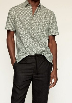 Mężczyzna w krótkim rękawie koszuli zapinanej na guziki z wzorem diamentu i czarnych spodniach z troczkami, z częściowo schowanymi rękami w kieszeniach.