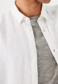 Camisa de lino blanca con un cuello abotonado, superpuesta sobre una camiseta de rayas grises. Presenta pequeños botones y un ajuste relajado.