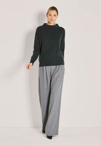 Frau in einem dunkelgrünen Langarmpullover, grauen weit geschnittenen Hosen, schwarzen High Heels und langen silbernen Ohrringen, die vor einem einfarbigen Hintergrund steht.