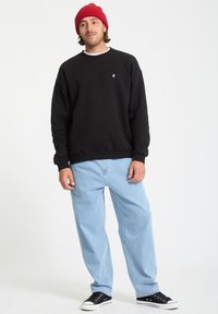 Sort sweatshirt med rund hals, lange ærmer og lille logo. Lyseblå vide jeans. Sorte sneakers med hvide gummisåler og rød beanie.