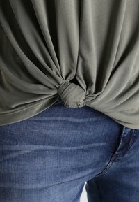 Olivgrünes geknotetes Oberteil aus weichem Stoff mit einem Drehdetails an der Taille, kombiniert mit blauen Denim-Jeans.