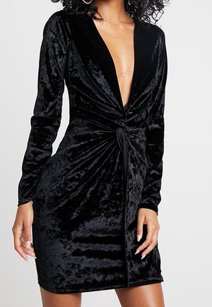 Woman wearing a black velvet long-sleeve mini dress with deep V-neck and front knot detail, standing against a plain background.