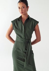 Abito verde con maniche corte a sbuffo, chiusura frontale con bottoni, vita legata e inserti laterali. Tessuto liscio e texture con una vestibilità sartoriale.