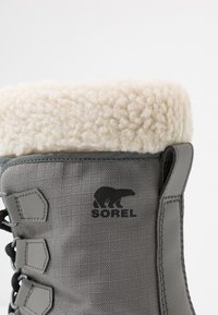 Botte imperméable grise avec un col en fausse fourrure blanche, tige texturée, logo noir et design à lacets pour un maintien sûr.