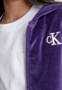 Fialová sametová zipová mikina s bílým logem "ck", nošena přes bílé tričko s kulatým výstřihem. Měkká textura a viditelný detail stříbrného zipu.