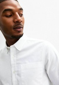 Camicia bianca con colletto, taschino singolo sul petto e tessuto texturizzato. Il modello indossa la camicia su uno sfondo neutro.
