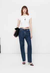 Witte cropped t-shirt met grafische print, blauwe high-waisted rechte jeans, zwarte clutch en zwarte hoge sandalen. Minimalistische stijl.