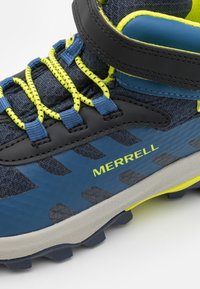 Zapato de senderismo azul con acentos en negro y amarillo, que presenta una parte superior texturizada, suela gruesa y un dibujo prominente para ofrecer tracción. Se ve el nombre de la marca.