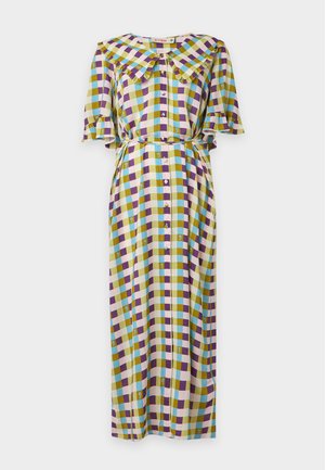 Femme portant une longue robe à carreaux multicolores avec un large col volanté et des ballerines noires, debout sur un fond uni.