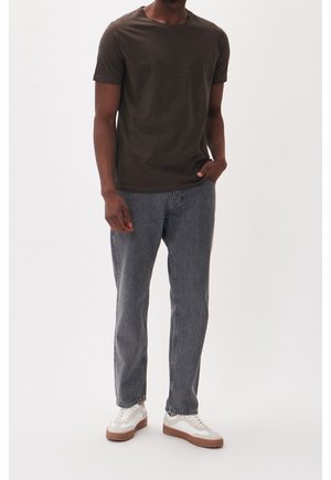 Mann trägt dunkelbraunes T-Shirt, graue Jeans und weiße Sneaker, steht mit einer Hand in der Tasche vor einfachem Hintergrund.