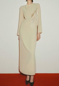 Femme en longue robe beige à manches longues avec un détail à franges sur la poitrine, portant des escarpins noirs à bout pointu, debout sur un sol rouge.