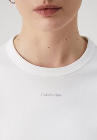 Bílý bavlněný tričko s žebrovaným kulatým výstřihem a decentním vyšitým logem "Calvin Klein" na hrudi. Hladká, jemná textura.
