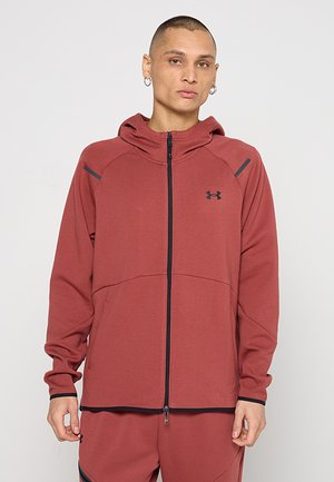 Persona che indossa una felpa con cappuccio con zip Under Armour color ruggine e pantaloni coordinati con cerniera nera e dettagli di rifinitura, in piedi davanti a uno sfondo semplice.