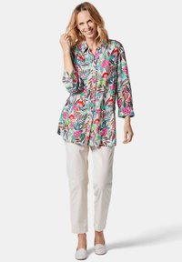 Blumenhemd in lebhaften Farben mit grünen Blättern, kombiniert mit weißen Hosen und hellen Mokassins.