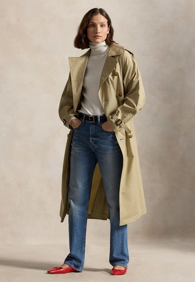 Γυναίκα που φοράει μπεζ παλτό τρεντσκότ πάνω από λευκό ζιβάγκο, μπλε τζιν με μαύρη ζώνη, και κόκκινα μυτερά φλατ, στέκεται με τα χέρια στις τσέπες.