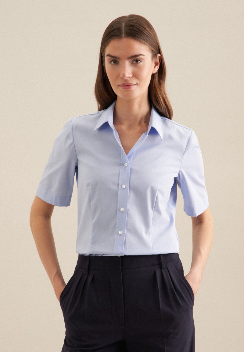 Camicia azzurra chiara con colletto e maniche corte, dotata di chiusura con bottoni, realizzata in tessuto morbido. Abbinata a pantaloni scuri plissettati.