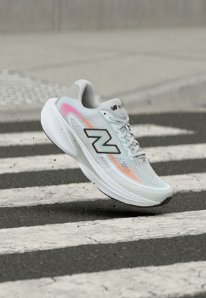 Valkoinen ja harmaa New Balance -juoksukenkä, jossa värikkäitä yksityiskohtia, ilmassa jalankulkijan suojatien yläpuolella asfaltilla.