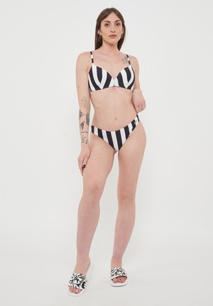 Bikini a righe bianche e nere con spalline regolabili. Presenta un top a triangolo e uno slip a vita bassa. Indossato con sandali con pattern.