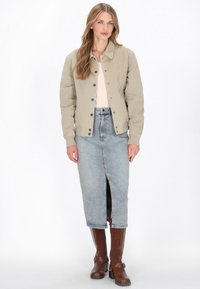 Beige vászon dzseki gombos zárással, világos színű felsővel és elöl hasított világos farmerral párosítva. A megjelenést barna térdig érő csizmák teszik teljessé.