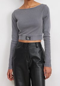 Grå ribbad långärmad croppad topp med bred halsringning, prydd med en svart CK Jeans-logotyp vid fållen, som är ihopparad med svarta high-waisted läderbyxor.